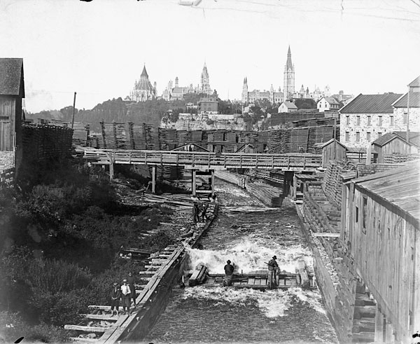 Une photo en noir et blanc qui montre deux personnes à bord un radeau sur la glissoire à bois de la Chaudière, avec les édifices du Parlement dans l'arrière-plan.